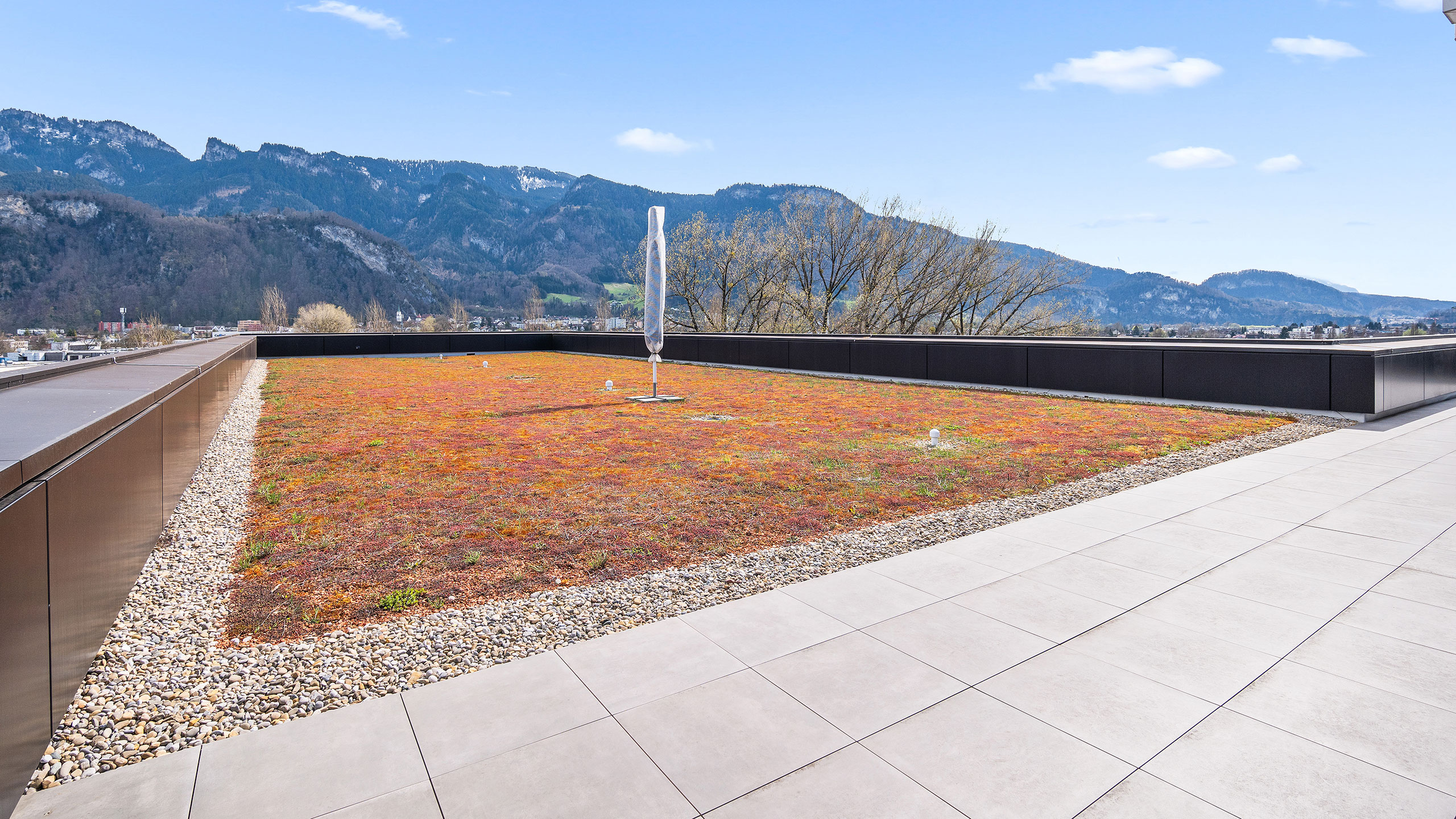 Das Bild zeigt die begehbare Grünfläche auf der Dachterrasse im Bürokomplex Schlatt1 in Hohenems/Österreich. Im Hintergrund ist das wunderschöne Bergpanorama der Alpen sichtbar.